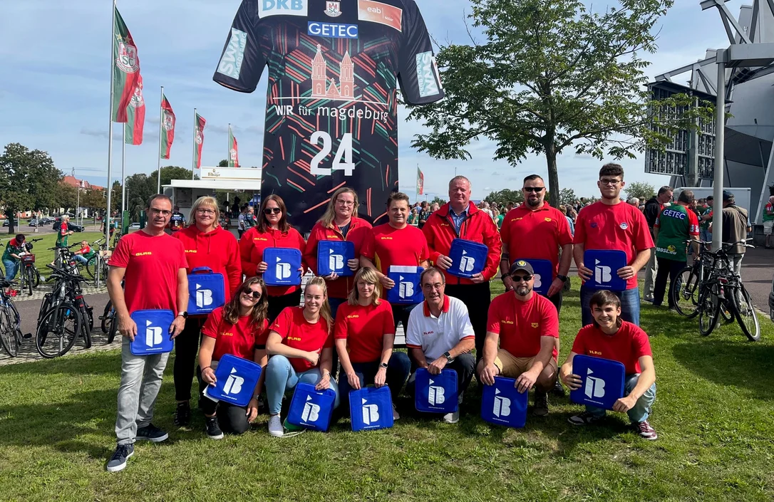 Eine Gruppe Menschen vom DLRG mit IB-Logos in der Hand vor einer Arena in Magdeburg