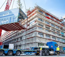 Eingerüsteter Neubau, davor ein Schwerlasttransporter und ein Kran, an dem ein mit Grünen Zweigen geschmückter Container hängt. (Bild: Universitätsklinikum Halle)