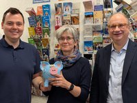 Drei Personen vor einer Wand mit Flyern. Die Frau in der Mitte hält ein Sparschwein.