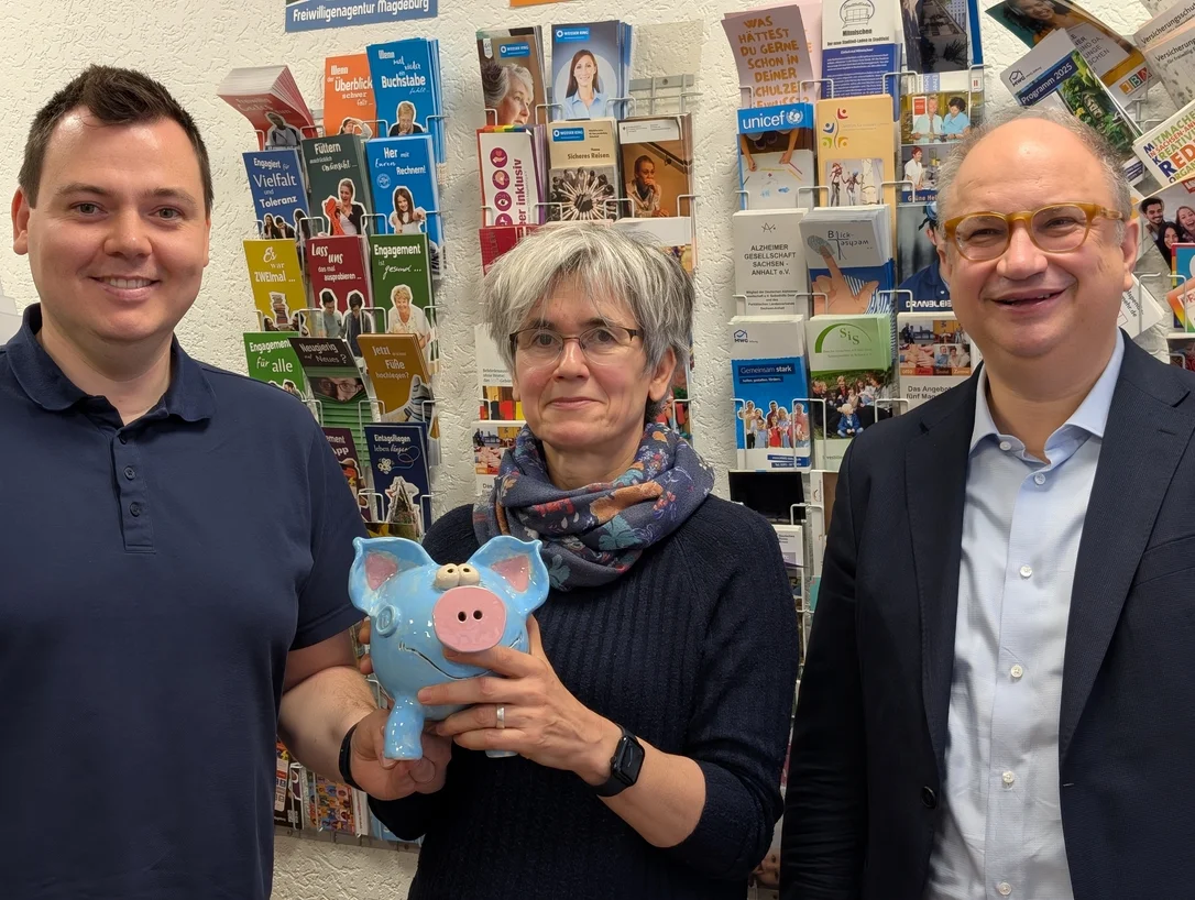 Drei Personen vor einer Wand mit Flyern. Die Frau in der Mitte hält ein Sparschwein.