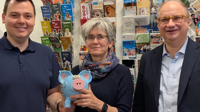 Drei Personen vor einer Wand mit Flyern. Die Frau in der Mitte hält ein Sparschwein.