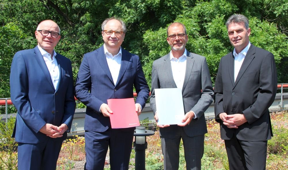 Auf dem Bild sieht man vier Herren mittleren Alters in Anzügen nebeneinander stehen. Die beiden mittleren Herren halten jeweils eine Dokumentenmappe in der Hand