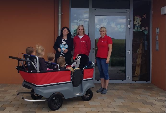 Auf dem Bild sieht man einen 6-sitzigen Krippenwagen, besetzt mit 5 Kindern und einem großen Plüsch-Maulwurf. Daneben stehen drei Frauen.