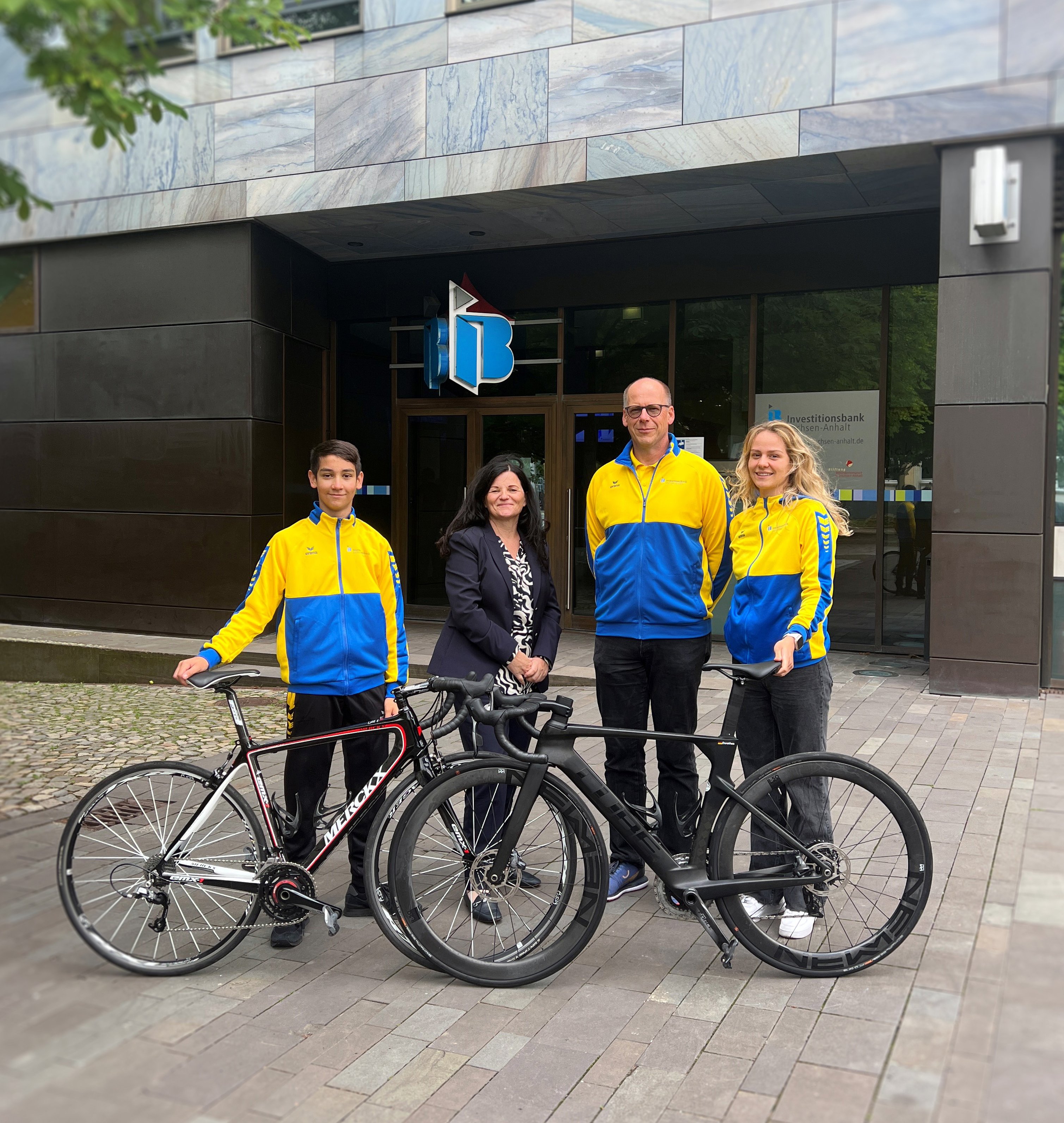 Auf dem Bild zu sehen sind zwei Fahrräder, dahinter stehen ein Mädchen und ein Junge sowie ein Mann und eine Frau. Im Hintergrund zu sehen ist der Haupteingang der IB mit dem Logo.