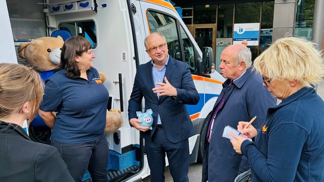 Gruppe von Menschen vor einem ASB-Wünschewagen im Gespräch, mit Plüschbär im Wagen und Hund am Boden. 