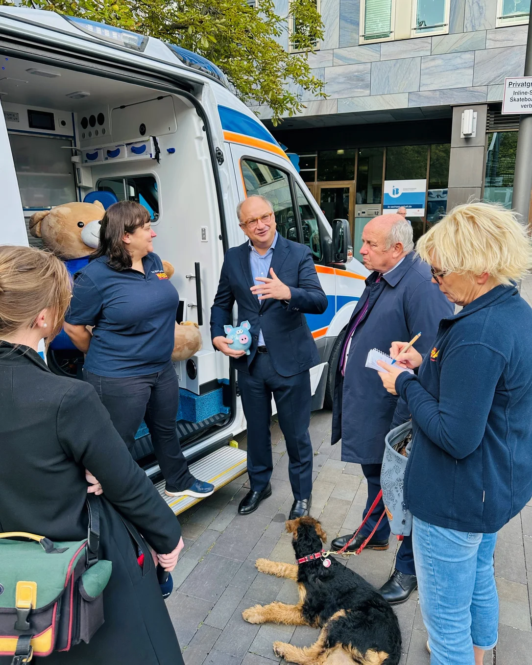 Gruppe von Menschen vor einem ASB-Wünschewagen im Gespräch, mit Plüschbär im Wagen und Hund am Boden. 