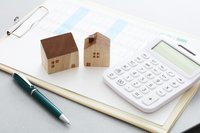 Ein Taschenrechner liegt auf einem Klemmbrett neben zwei kleinen hölzernen Hausfiguren