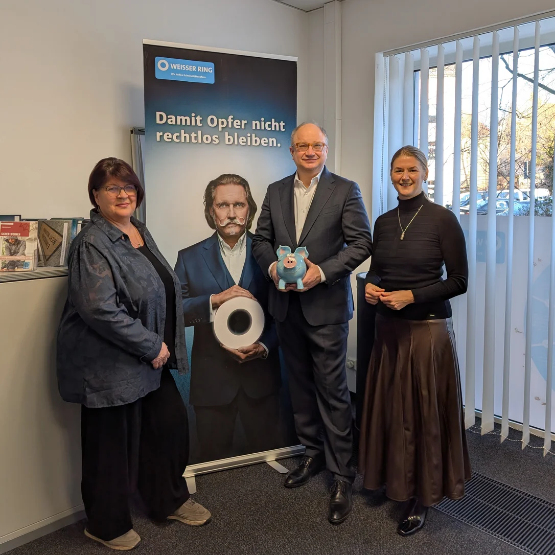 Drei Personen stehen vor einem WEISSER-Ring-Banner mit der Aufschrift "Damit Opfer nicht rechtlos bleiben".