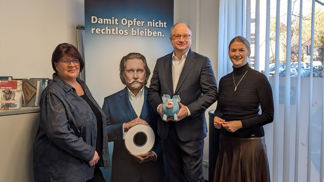 Drei Personen stehen vor einem WEISSER-Ring-Banner mit der Aufschrift "Damit Opfer nicht rechtlos bleiben".