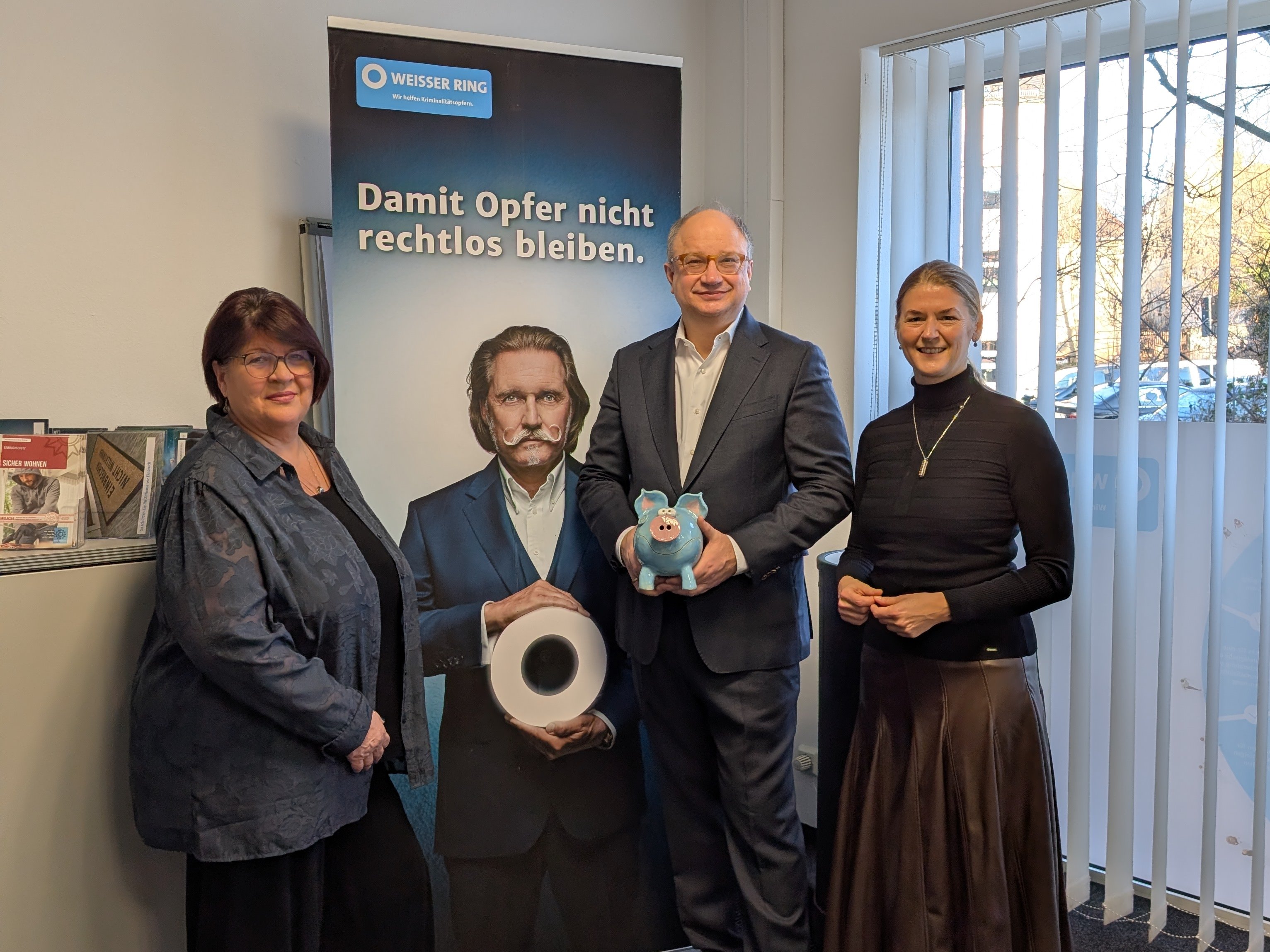Drei Personen stehen vor einem WEISSER-Ring-Banner mit der Aufschrift "Damit Opfer nicht rechtlos bleiben".