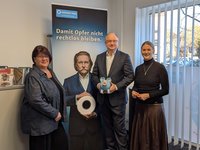 Drei Personen stehen vor einem WEISSER-Ring-Banner mit der Aufschrift "Damit Opfer nicht rechtlos bleiben".